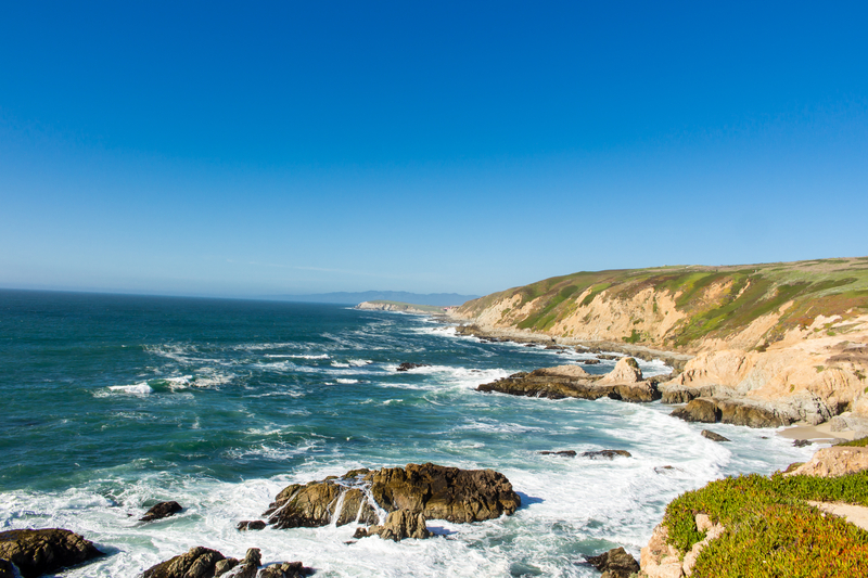 Grassy tan cliffs overlooking crashing waves and turquoise ocean on a sunny day.